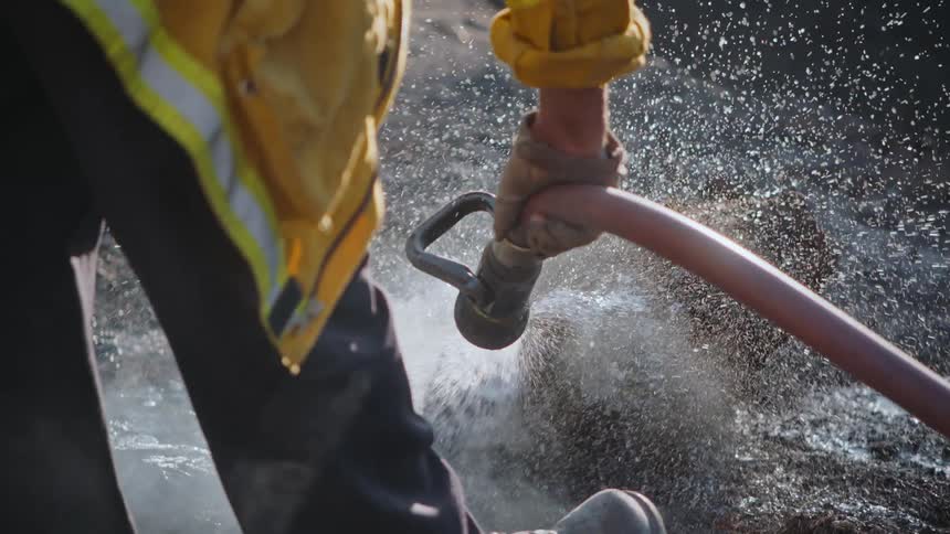 A fireman is standing outside holding a hose as he sprays water on a burning log and smoke rises