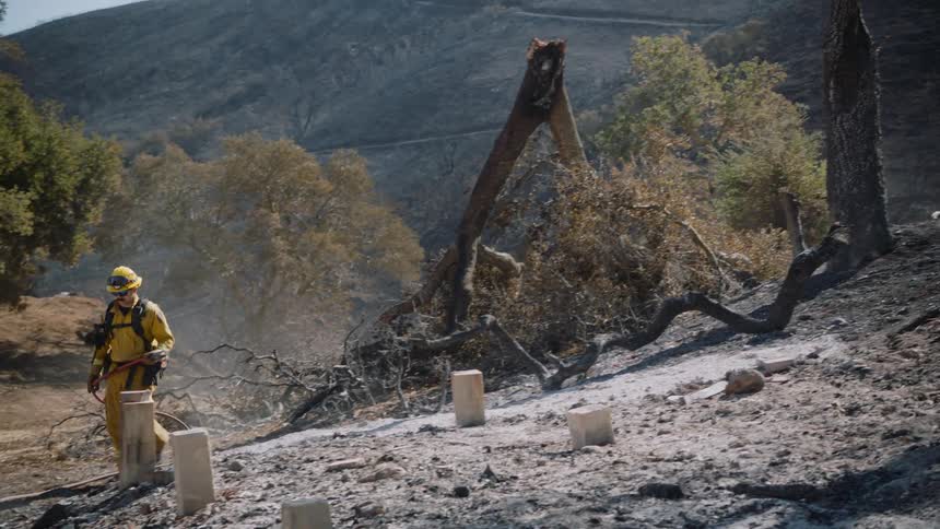 Firefighter sprays burnt ground with charred slope in the background