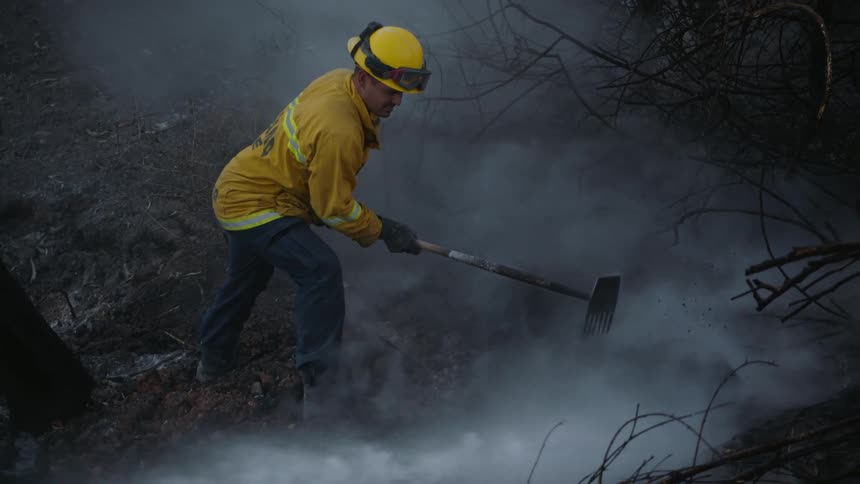 A firefighter rakes the ground while a fire hose soaks the land