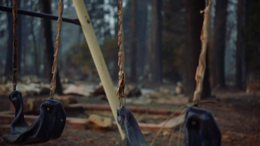 Melted swings sway on swing set in burnt area in conifer forest