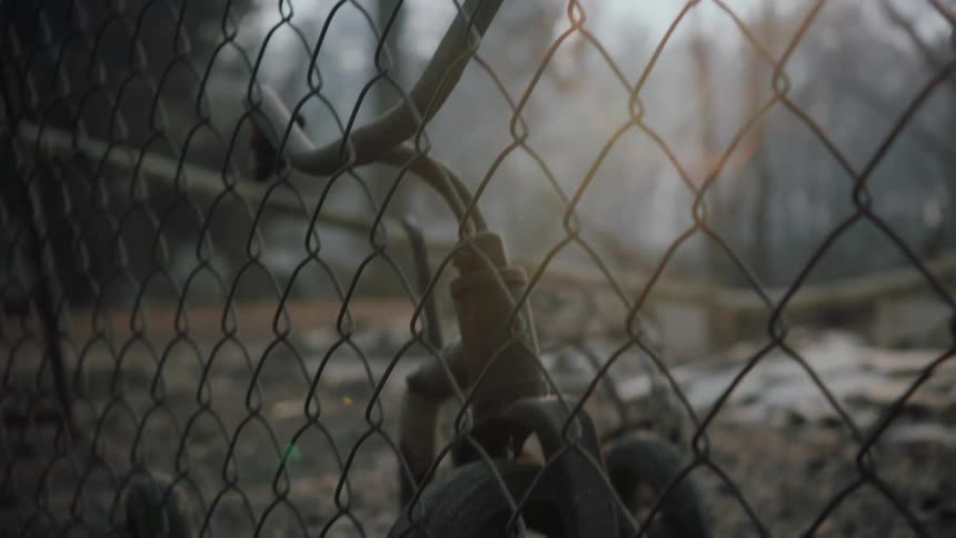 A charred and burned tricycle sits among debris near a chain link fence and there are woods in the distance in a smokey sky