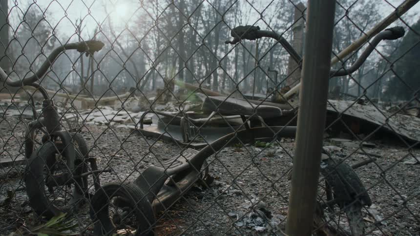 Burnedout tricycles and the rubble of a collapsed building rest behind a metal fence near a forest
