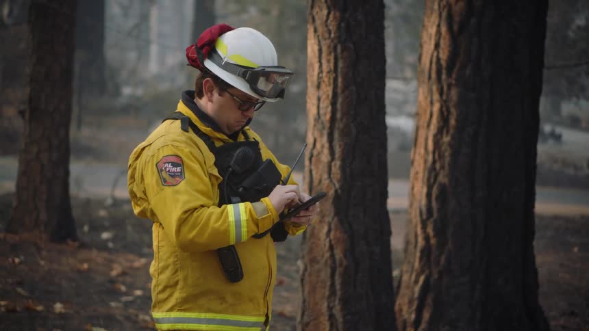 A fireman is standing outside near trees using his cell phone and there is smoke in the distance