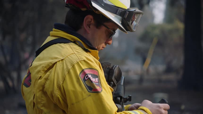 A fireman is standing outside near trees using his cell phone and there is smoke in the distance