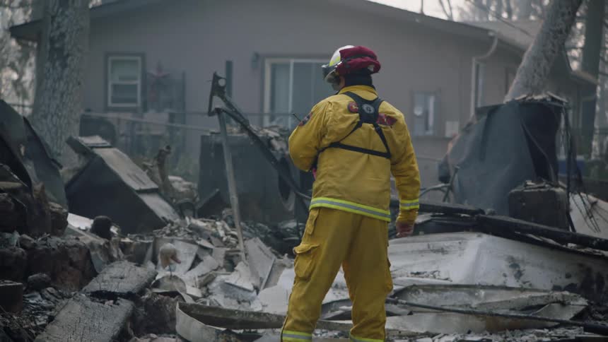 A firefighter in a yellow jumpsuit climbs the steps in front of the rubble of burnedout homes and trees