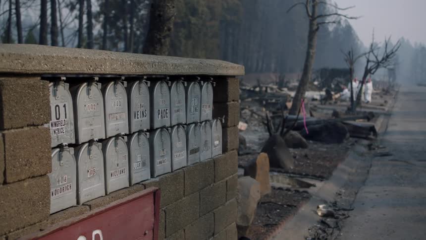 A bunch of wornout metal mailboxes stand before a ruined neighborhood street while a crew in white works in the background