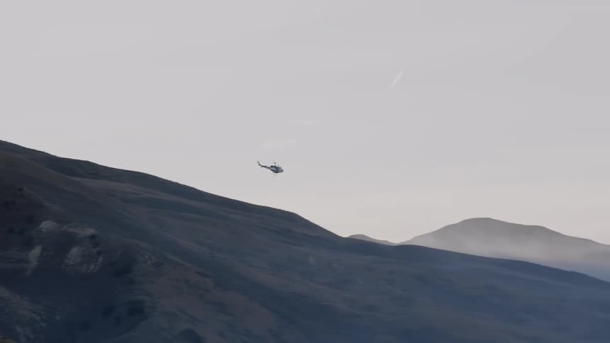 A helicopter flies over a mountain ridge covered in grass on a foggy day