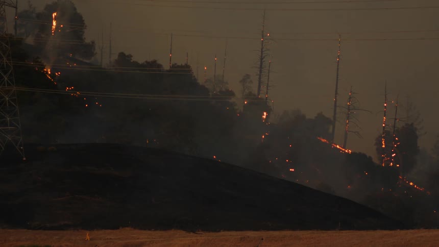 Person wearing firefighting uniform walks through field with charred hills and trees still glowing with embers in the background