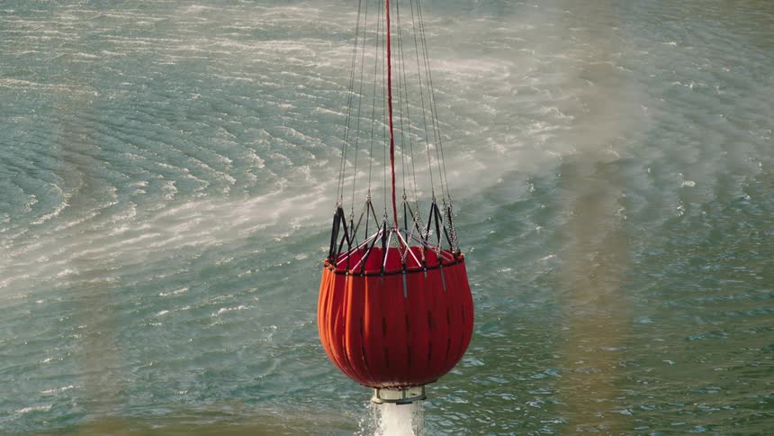 A red helicopter bucket is being refilled and lifted out of a large reservoir