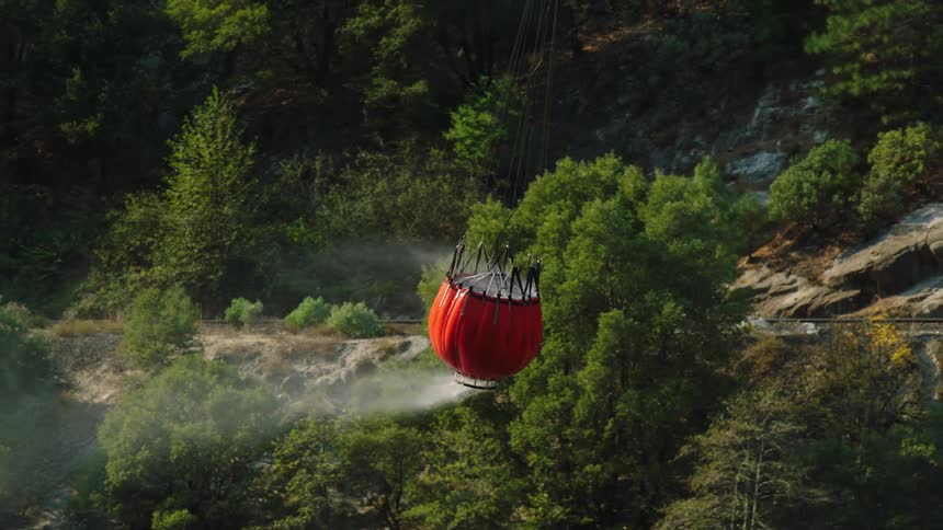 Red helicopter bucket spills water while moving over snow and conifer covered mountain side
