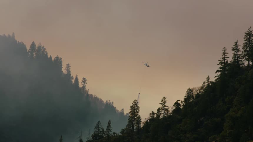 A helicopter flies over forested mountains in a smoke filled sky carrying water to put out a forest fire