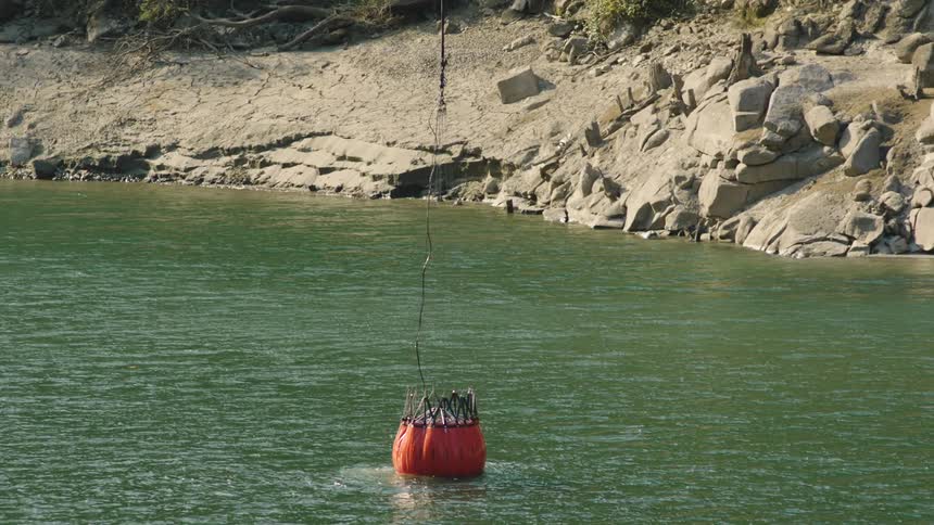 Red helicopter bucket leaks water while it is lifted from green lake water near boulder strewn shore