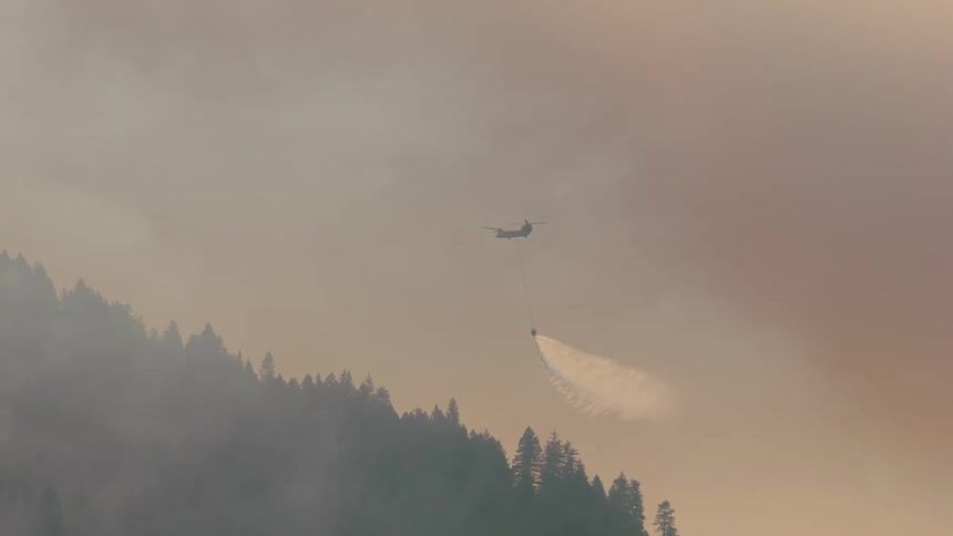 Firefighting helicopter flies through smoky air and spreads water from bucket onto conifer forest