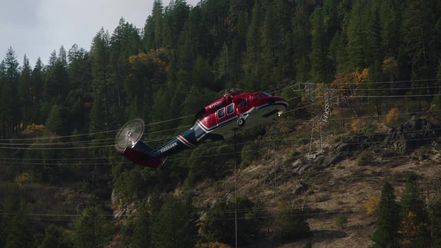 A helicopter carrying a specialized bucked suspended on a cable collects water for aerial firefighting