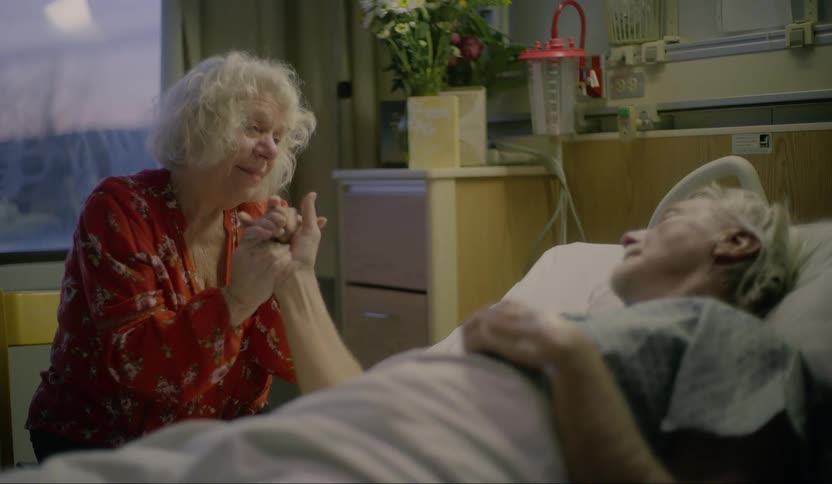 A woman holds her husbands hand as he lies in a hospital bed