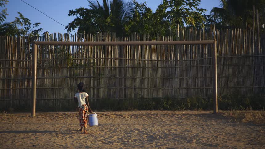 Child carrying a bucket stops to look at a soccer goal on a tropical country