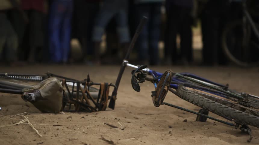 Two bicycles laying on the dirt ground as a crowd of people are gathered feet away