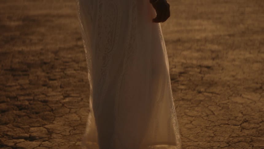 Woman wearing a white dress walks barefoot on the desert at daytime
