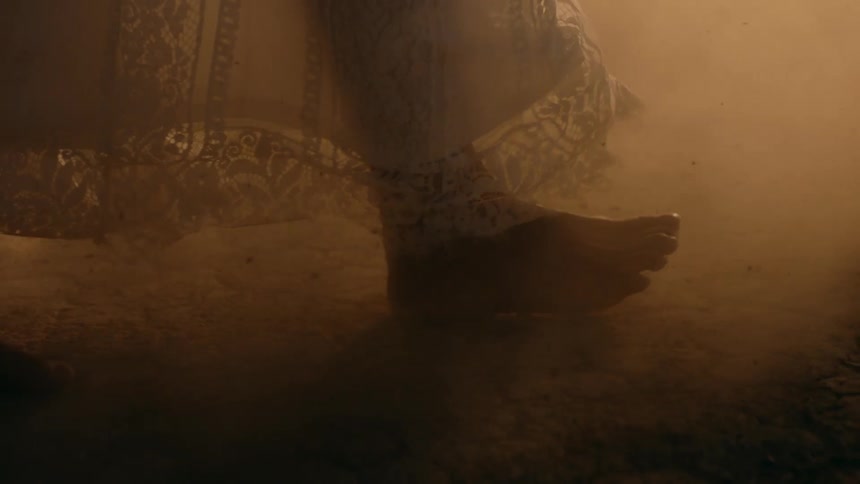 A persons feet walking barefoot on dry desert ground wearing a white gown