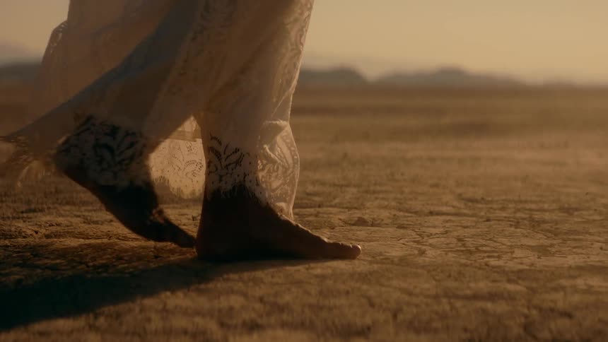 Dust blows around her feet as woman wearing lace walks across ground