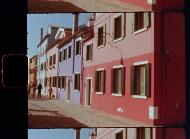 The setting sun shines onto a small alley or street with colourful house facades and a family of three people including a child walking or strolling in a small town on a beautiful evening caught on vintage film