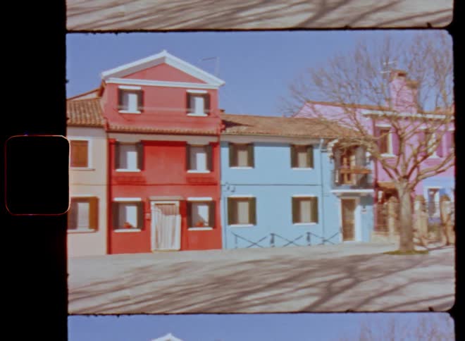 Panning vintage clip of colorful houses or rowhouses standing next to a small street or road with a bare and leafless tree in the middle of the town square as the sun shines with a clear blue sky