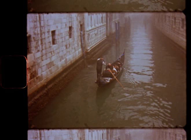 A group of people are being rowed in a canoe or Venetian gondola down the canal with brick stone walls and reflections on the surface of the water enjoying the boat ride on a foggy day on vintage film footage