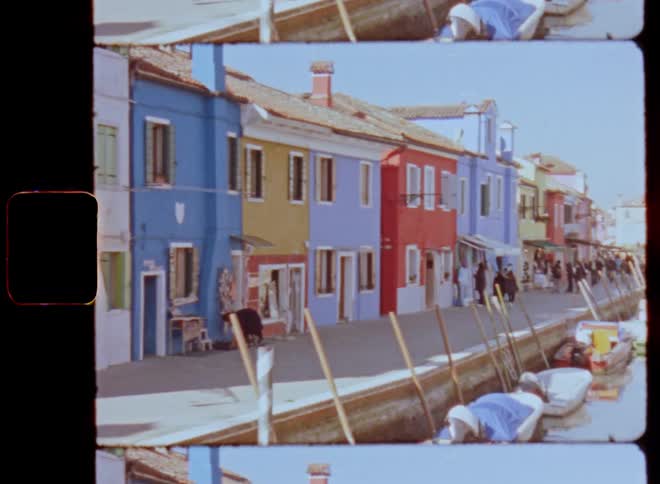 Colorful facades of houses near the canals in Venice as people or groups of tourists walk or stroll along the pier next to parked motor and rowing boats on a sunny springtime day caught on vintage film