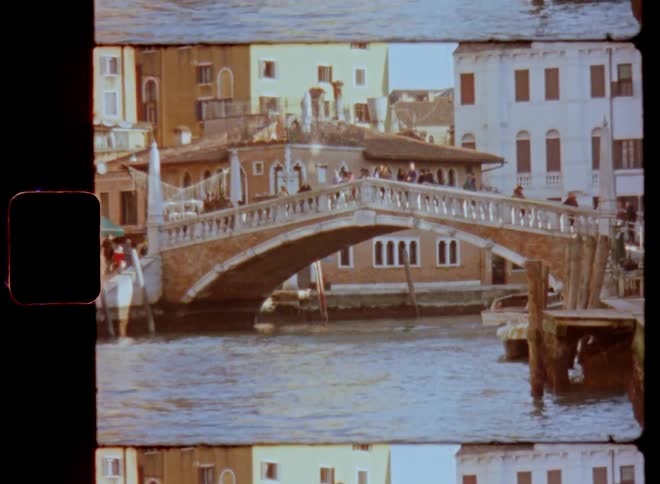 Many pedestrians or tourists walk over a crowded bridge going over a waterway or canal between buildings as the sun shines and people enjoy their holidays while traveling through Italy and Europe