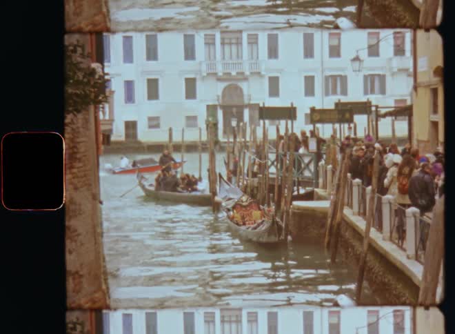 A crowd of people standing in line waiting on a sidewalk next to water with parked boats floating next to the pier on a harbor or port surrounded by waterfront buildings on vintage film