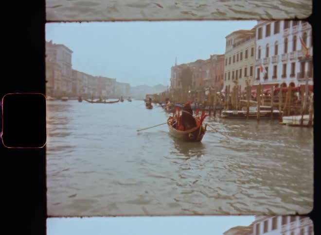A standing man and other people row a gondola on the large Venice canals while other boats drive or ride over the river surrounded by large buildings and old houses caught on vintage film