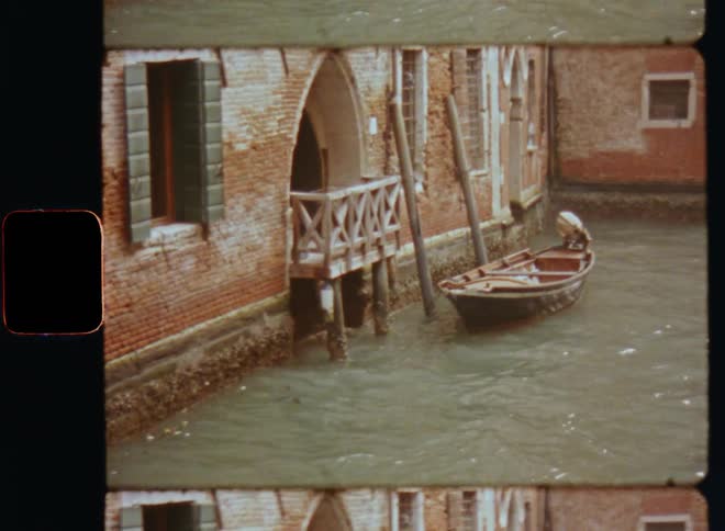 Vintage video of an old parked motorboat floating next to an old house or brick building with a porch or balcony in a city filled with waterways such as rivers and canals