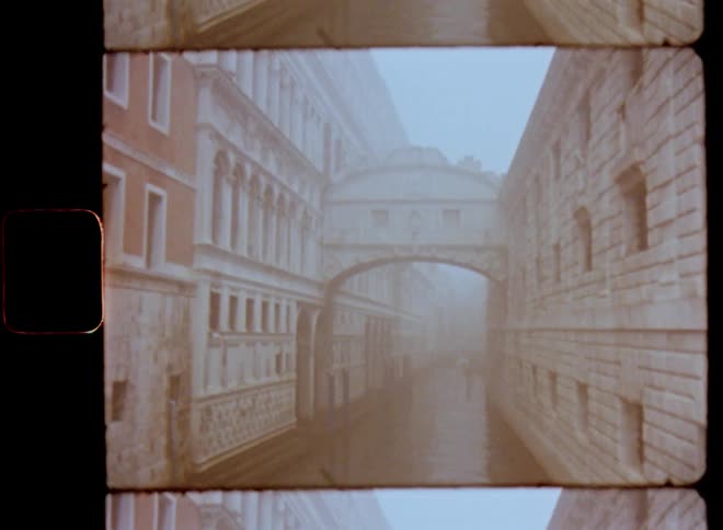 Vintage footage of water canals going through old brick buildings with many windows looking onto a high bridge connecting two stone walls on a foggy overcast day with gray sky and gloomy light