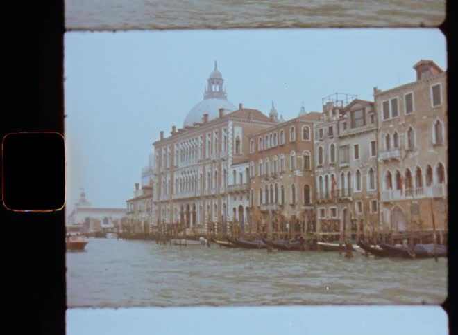 Driving on a ship looking over the water onto the waterfront buildings and houses with gondolas and other boats parked at the pier or shore and a large cathedral in the background of the city