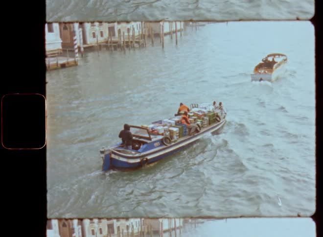 Handheld Super 8mm film footage of two motorboats transporting people while driving on a large river or wide canal in Venice with old majestic buildings and a large cathedral standing at the shore or edge of the waterway