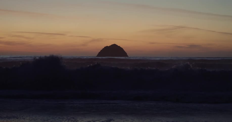 Waves splash as they roll into shore as the silhouette of a rock stands on the horizon in an orange sunset sky