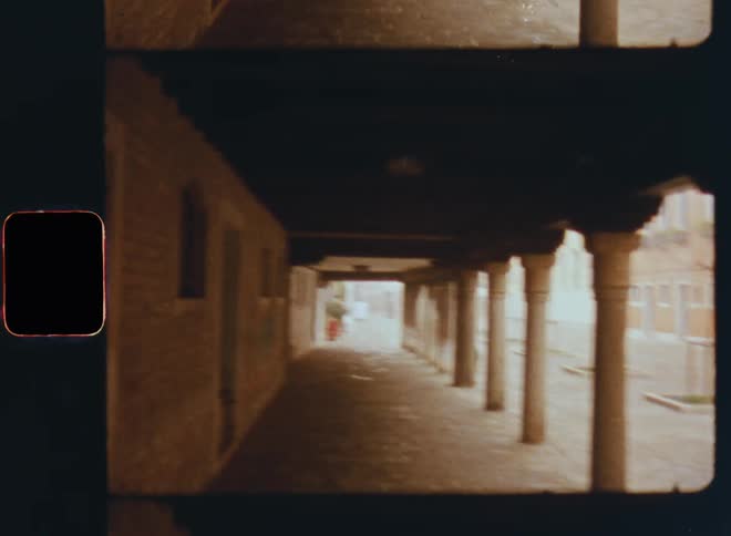 Walking through a corridor outdoors underneath a porch or stem with pillars or columns on the right and windows and doors on the left side as the roof of the old building creates shadow at the entrance of the house