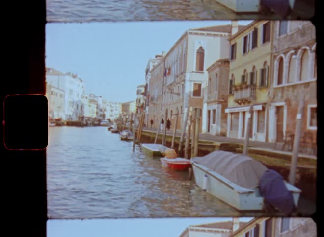 Floating or driving a boat on a river surrounded by piers and old buildings in a Mediterranean city as small motor boats are parked at the edge of the waterfront houses and pedestrians move alongside the canal