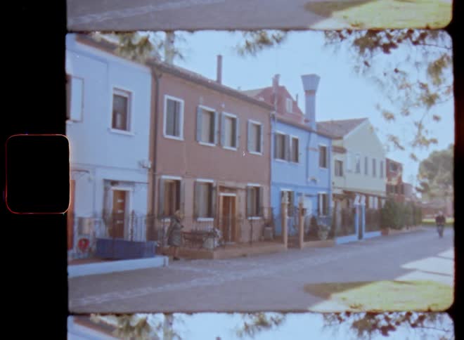 Panning Super 8 shot of people moving along street or road with row houses painted in various colourful colors with lots of windows and entrance doors