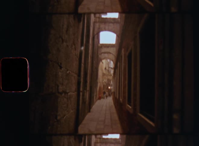 Walking on a dark and narrow arched alley between tall buildings on an ancient city as windows and doors in the stone walls pass by caught on vintage S8 film with shaky camera movement
