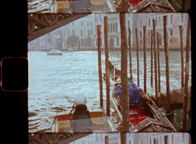 Old gondolas and a motorboat parked on a pier underneath a bridge tied up to wooden poles as other boats pass by on the canal or river in Venice caught on vintage Super 8 film