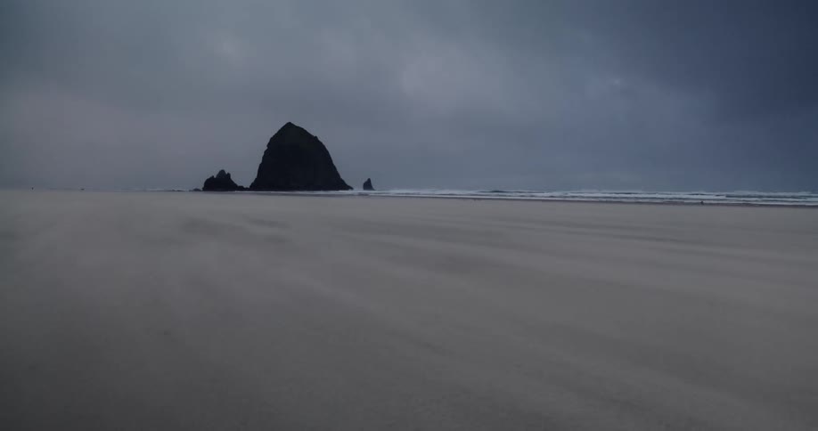 Sand blows across a beach in front of a large rock outcropping