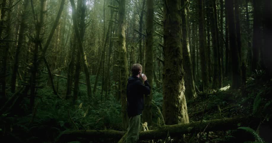 A man is standing near trees in the forest holding an audio recorder in his hand as he speaks