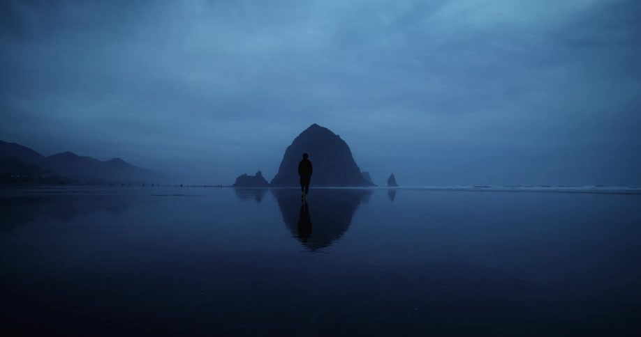 A man is walking alone along a beach shoreline with cloudy skies at dusk