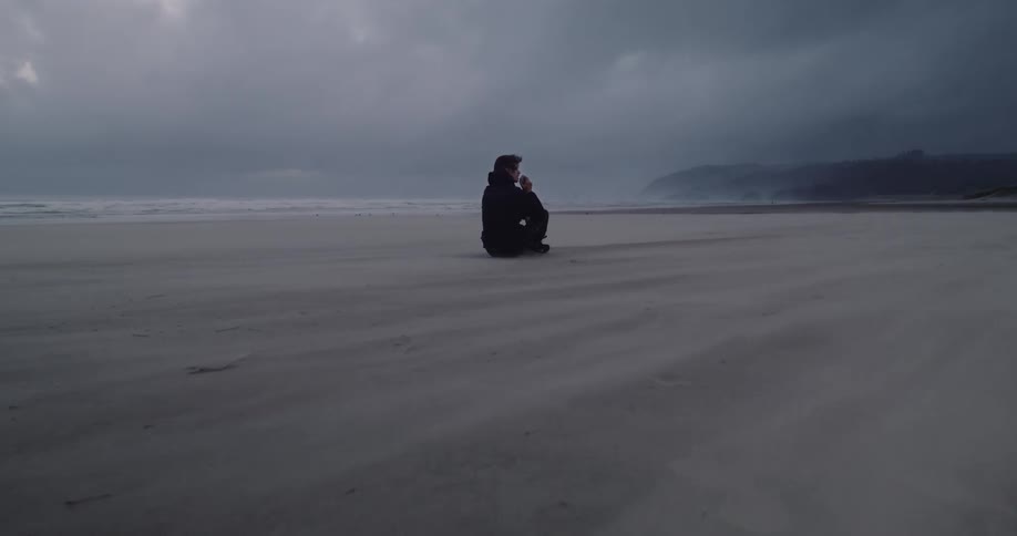 Wind blows sand across beach where person sits under an overcast sky