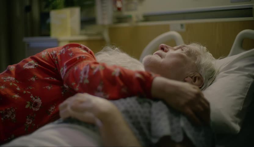 An elderly woman hugs her husband tight lying in a hospital bed