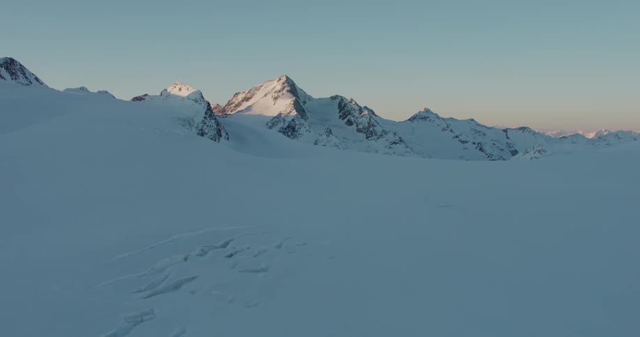 A snow covered landscape gives rise to a rocky mountain range in the distance