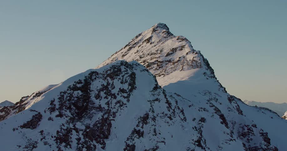 The sun shines on a snow covered mountain ridge with a peak high in the clear sky