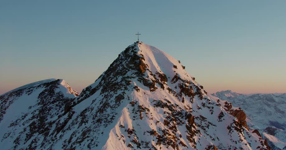 A cross stands on a snow covered mountain peak surrounded by mountains as the sun shines in a clear sky