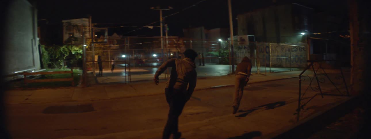 Two men run down an alley and hop a fence at night